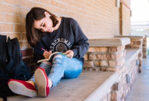 Brown Haired Caucasian teen girl wearing gray sweatshirt, torn jeans, and airpods, sitting on concrete bench writing in book. 